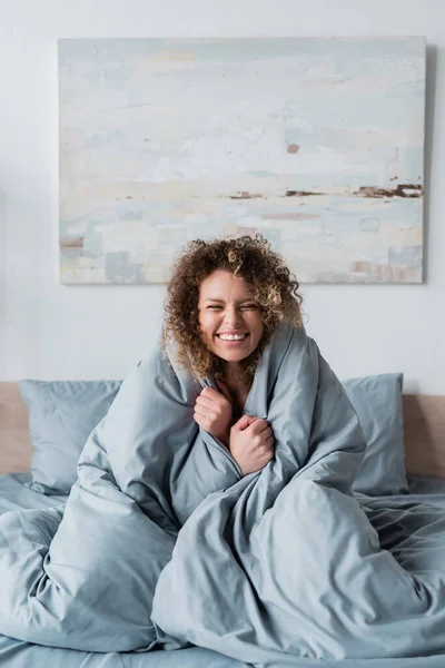 Возбужденная и кудрявая женщина с закрытыми глазами, сидящая на кровати под серым одеялом — стоковое фото