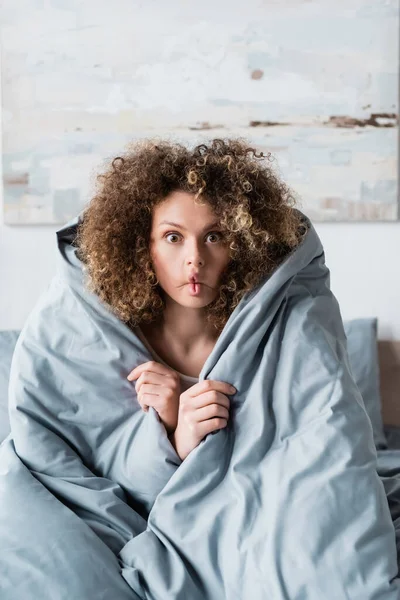 Кудрявая женщина со смешной гримасой, завернувшаяся в серое одеяло и смотрящая в камеру — стоковое фото