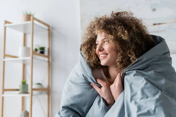 Веселая кудрявая женщина, завернутая в серое одеяло и смотрящая в спальню — стоковое фото