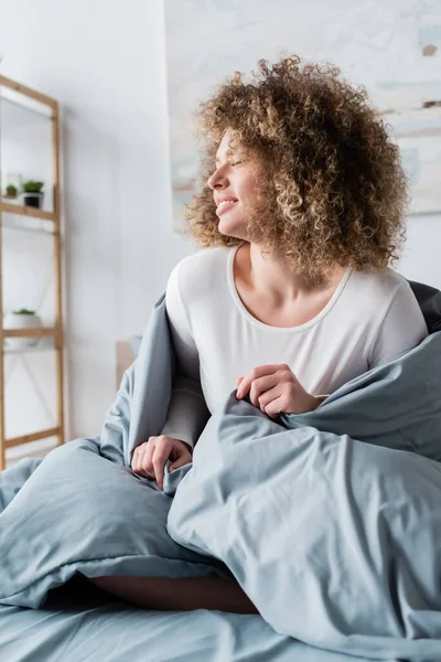 Freudige Frau mit welligem Haar sitzt unter grauer Decke und lächelt im Schlafzimmer — Stockfoto