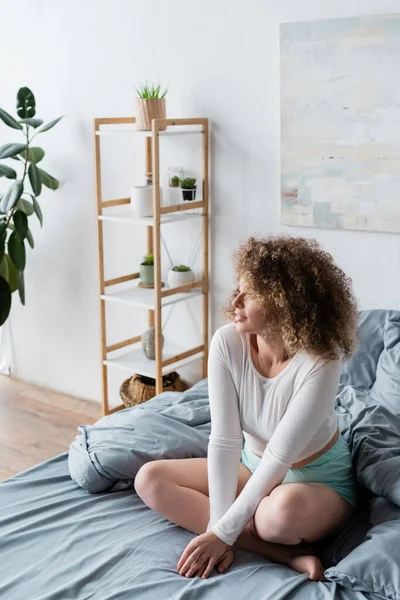 Femme bouclée assise sur le lit avec les jambes croisées près de rack flou avec des pots de fleurs — Photo de stock