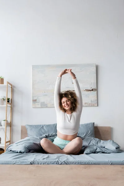Счастливая женщина, сидящая на кровати со скрещенными ногами и вытянутыми руками — стоковое фото