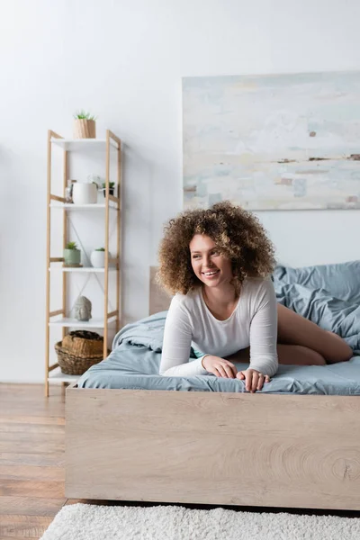 Jovem mulher encaracolado sorrindo na cama perto de cremalheira borrada com plantas em vaso — Fotografia de Stock