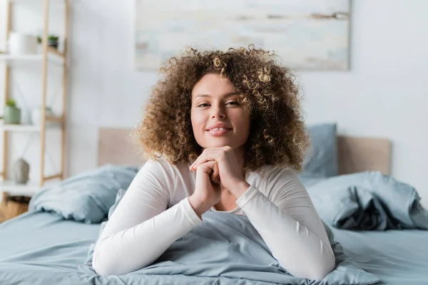 Pleased curly woman smiling at camera while lying on bed — Stock Photo