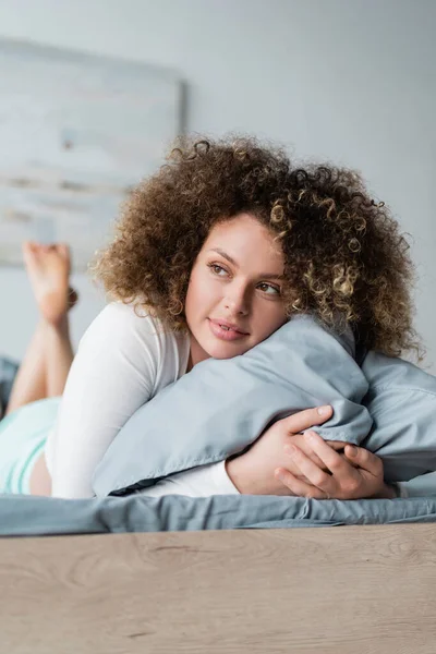 Feliz jovem mulher olhando embora enquanto abraçando travesseiro na cama — Fotografia de Stock