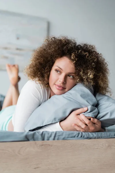 Heureuse jeune femme regardant loin tout en étreignant oreiller sur le lit — Photo de stock