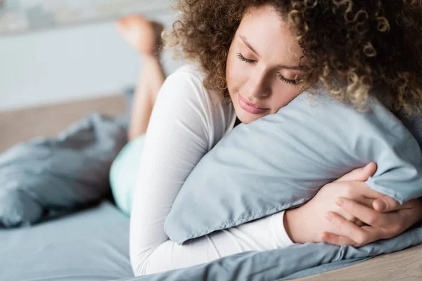 Zufriedene Frau umarmt Kissen, während sie mit geschlossenen Augen auf dem Bett liegt — Stockfoto