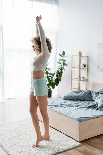 Longitud completa de mujer descalza en pijama blanco estirándose con las manos levantadas en el dormitorio - foto de stock