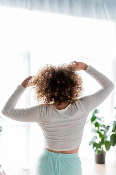 Vista posterior de la mujer joven con el pelo ondulado que estira los brazos en la mañana en casa - foto de stock
