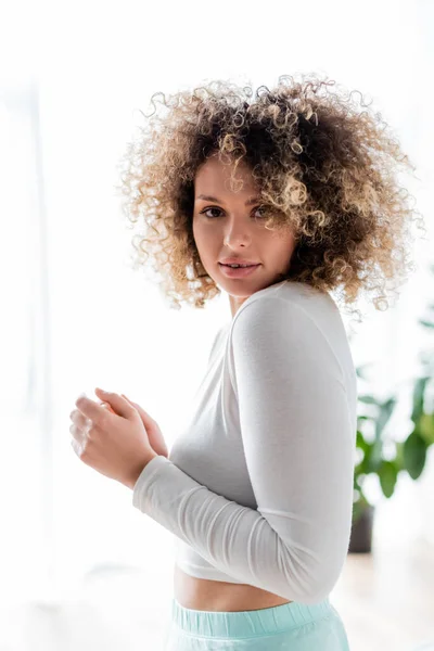 Pretty and curly woman in white home wear looking at camera at home — Stock Photo