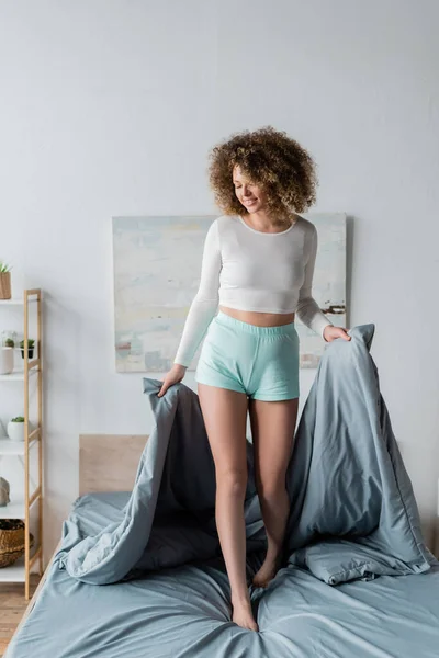 Повна довжина кучерявої жінки в білій піжамі тримає сіру ковдру, стоячи на ліжку — стокове фото