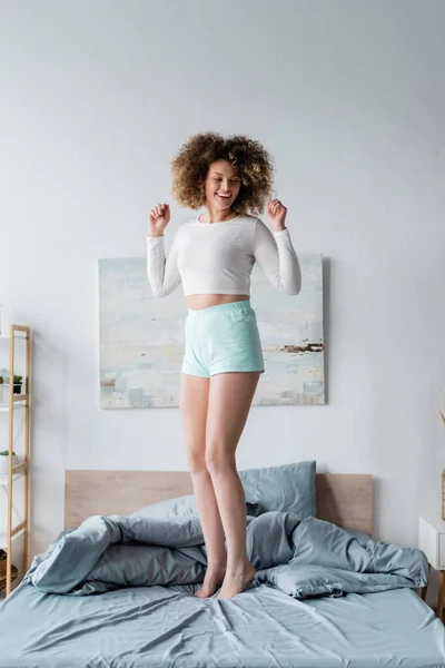 Pleine longueur de femme bouclée excitée en pyjama blanc debout sur le lit à la maison — Photo de stock