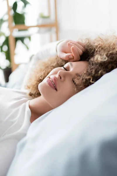 Jeune femme bouclée souriant tout en dormant sur le lit le matin — Photo de stock