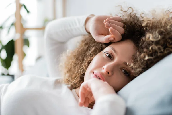 Affascinante e riccia donna con le mani vicino al viso guardando la fotocamera in camera da letto — Foto stock