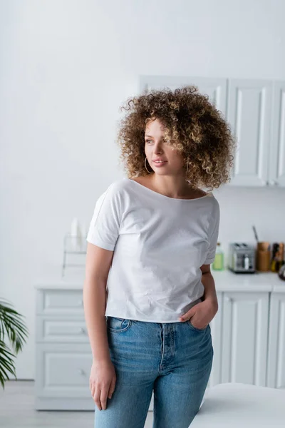 Mulher sorridente com cabelo ondulado em pé com a mão no bolso de jeans na cozinha — Fotografia de Stock