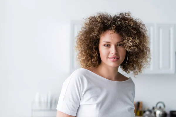 Curly woman in white t-shirt smiling at camera in kitchen — Stock Photo
