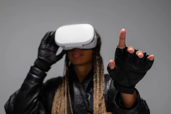 Розмита афріканська жінка в віртуальній реальності навушники і рукавички вказуючи пальцем ізольований на сірому — стокове фото
