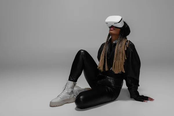 Afroamerikanerin in Lederjacke und Vr-Headset sitzt auf grauem Hintergrund — Stockfoto