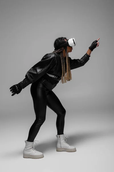 Side view of african american woman in virtual reality headset and leather jacket gaming on grey background — Stock Photo