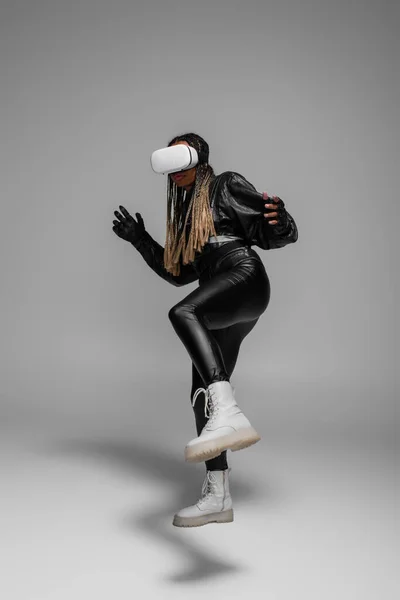 Elegante mujer afroamericana en chaqueta de cuero con auriculares vr sobre fondo gris - foto de stock