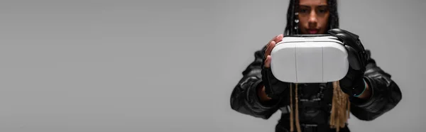 Mujer afroamericana borrosa en chaqueta de cuero con auriculares vr aislados en gris, pancarta - foto de stock