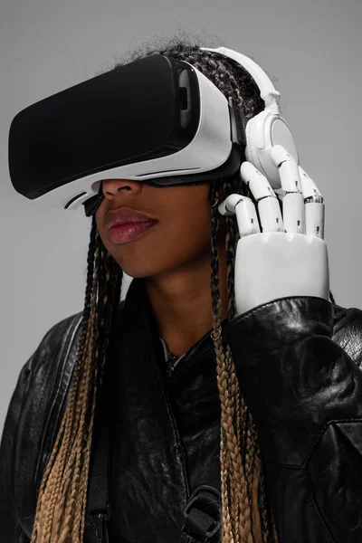 Африканская женщина с роботизированной рукой и VR-гарнитурой слушает музыку в VR-гарнитуре, изолированной на сером фоне — стоковое фото