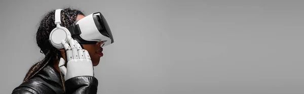 Vue latérale de la femme afro-américaine avec casque, casque vr et main robotique isolé sur gris, bannière — Photo de stock