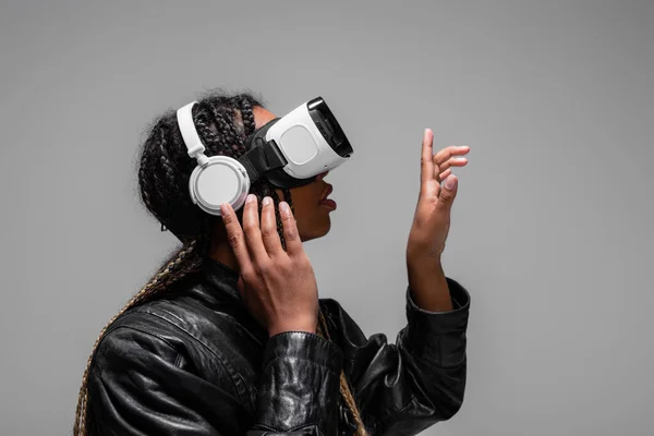 Side view of african american woman in headphones and vr headset pointing with finger isolated on grey — Stock Photo