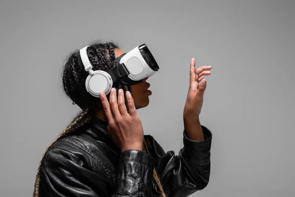 Vue latérale de la femme afro-américaine dans les écouteurs et le casque vr pointant avec le doigt isolé sur gris — Photo de stock