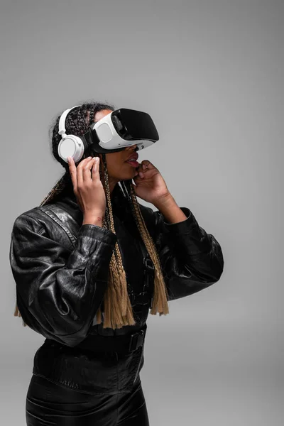 Femme afro-américaine en vr casque et veste en cuir écouter de la musique dans les écouteurs isolés sur gris — Photo de stock