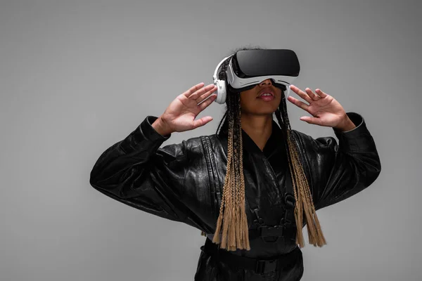 Femme afro-américaine élégante dans les écouteurs en utilisant un casque vr isolé sur gris — Photo de stock