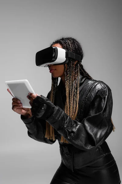 Африканская женщина в кожаной куртке и VR-гарнитуре с цифровым планшетом, изолированным на сером фоне — стоковое фото