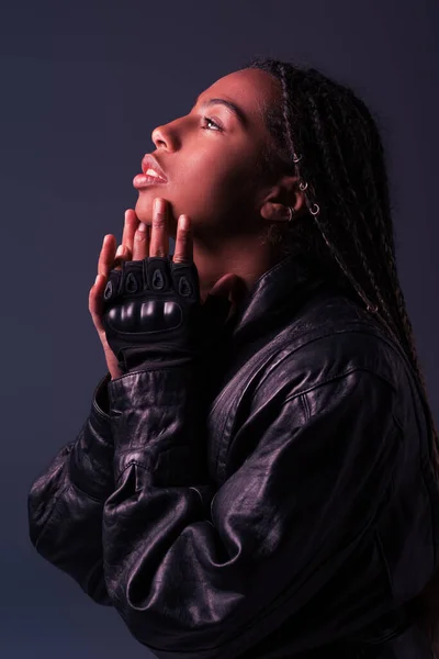 Vista lateral de la elegante mujer afroamericana en chaqueta de cuero y guante mirando hacia otro lado aislado en gris oscuro - foto de stock
