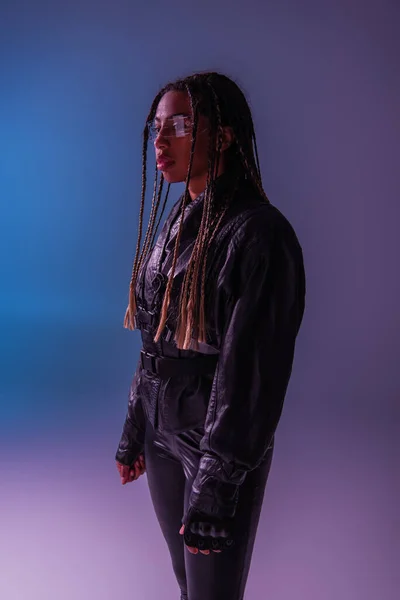 Mujer afroamericana con estilo en chaqueta de cuero y gafas inteligentes de pie sobre fondo azul y púrpura - foto de stock