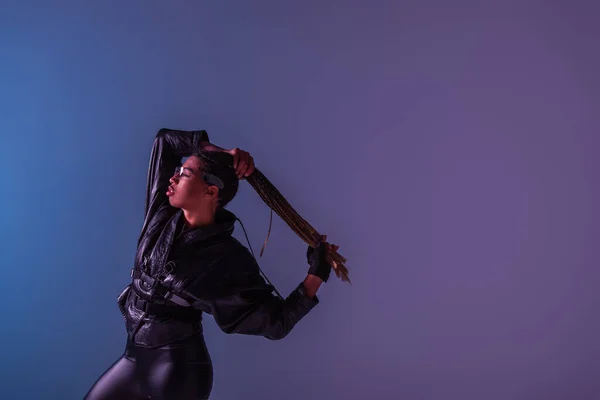 Vista lateral de la elegante mujer afroamericana en gafas inteligentes con coletas sobre fondo azul y púrpura - foto de stock