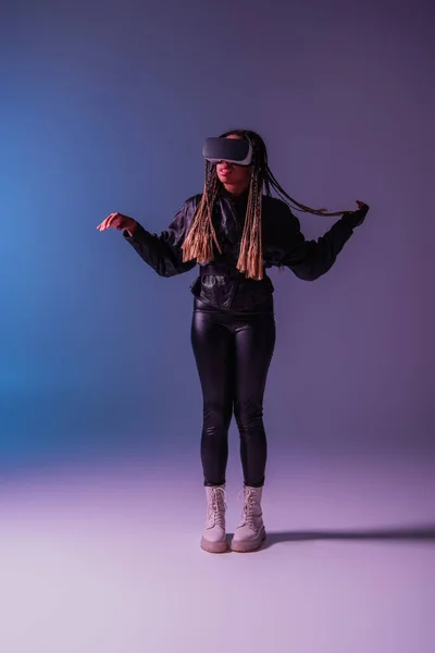 Pleine longueur de femme afro-américaine à la mode utilisant un casque vr sur fond bleu et violet — Photo de stock