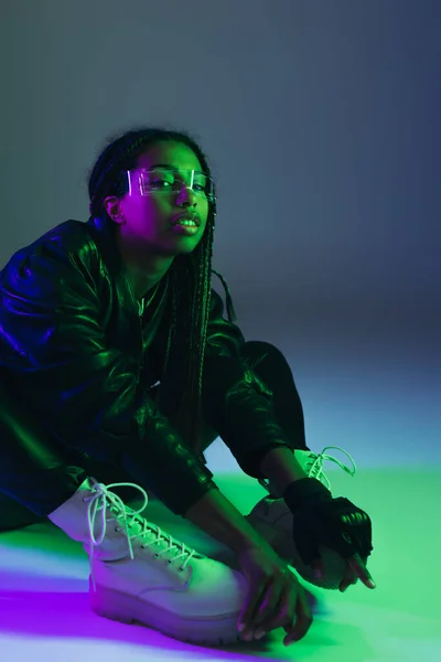 Mujer afroamericana de moda en gafas inteligentes sentadas sobre fondo azul con iluminación - foto de stock