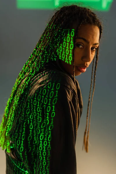 Stylish african american woman with projection of binary code looking at camera on grey background — Stock Photo