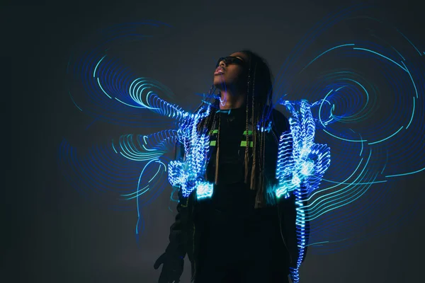 Elegante mujer afroamericana en gafas inteligentes de pie cerca de proyección abstracta sobre fondo gris - foto de stock