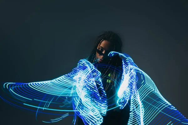 Mujer afroamericana en gafas inteligentes mirando la cámara cerca de luces de neón de abstracción sobre fondo gris - foto de stock