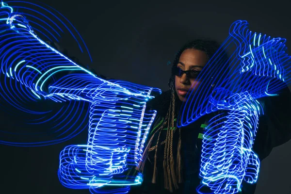 Mujer afroamericana en gafas inteligentes mirando hacia otro lado cerca de luces de neón sobre fondo gris - foto de stock