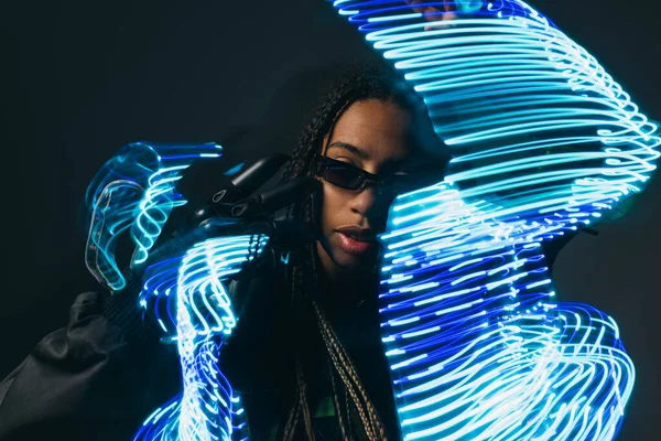 Mujer afroamericana con estilo en gafas inteligentes posando cerca de luces de neón abstractas sobre fondo gris - foto de stock