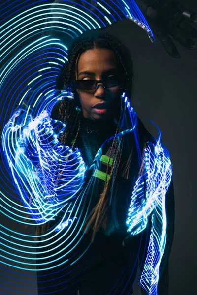 Longue exposition de la femme afro-américaine à la mode dans des lunettes intelligentes debout près sur fond gris — Photo de stock