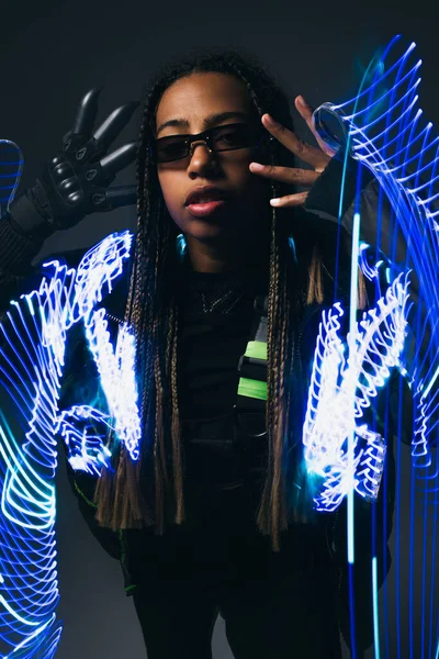 Afroamerikanerin in Handschuhen und smarter Brille steht neben abstrakten Neonlichtern auf grauem Hintergrund — Stockfoto