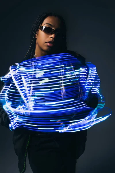 Femme afro-américaine élégante dans des lunettes intelligentes posant près de lignes néon sur fond gris — Photo de stock