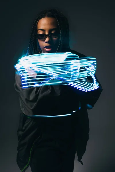 Stylish african american woman in smart glasses looking at neon lights on grey background — Stock Photo