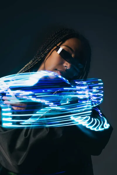 Retrato de mulher americana africana elegante em óculos inteligentes posando perto de luzes de néon azul no fundo cinza — Fotografia de Stock