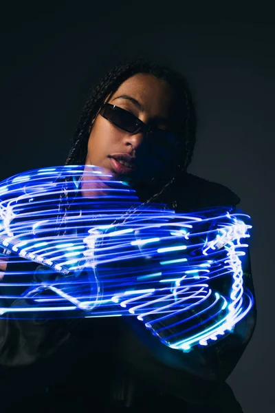 Femme afro-américaine branchée dans des lunettes intelligentes posant près de lumières bleues abstraites sur fond gris — Photo de stock