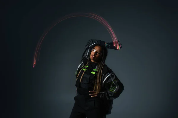 Larga exposición de la mujer afroamericana en gafas inteligentes posando cerca de la luz sobre fondo gris - foto de stock