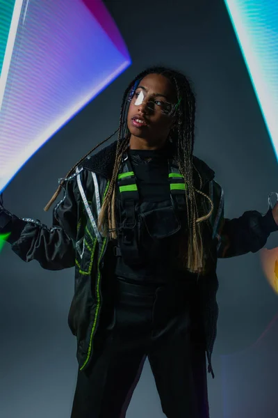 Mujer afroamericana en gafas inteligentes mirando hacia otro lado cerca de luces de neón sobre fondo gris - foto de stock